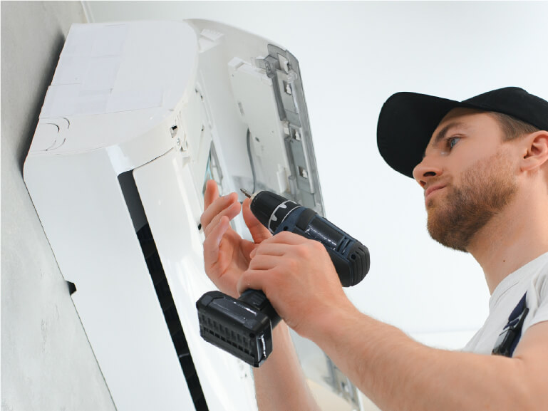 A man wearing a cap uses a power drill to install or repair a wall-mounted air conditioning unit.